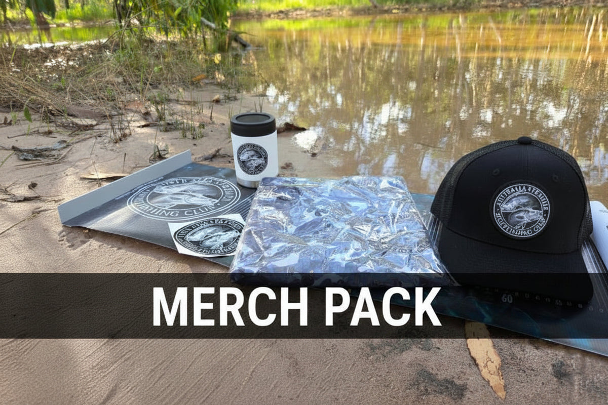 A black baseball cap with a circular 'Wilderness Fishing Club' logo sits next to a folded camouflage-patterned shirt, a white tumbler, and a branded sticker on a mat on a dirt path with a river in the background.